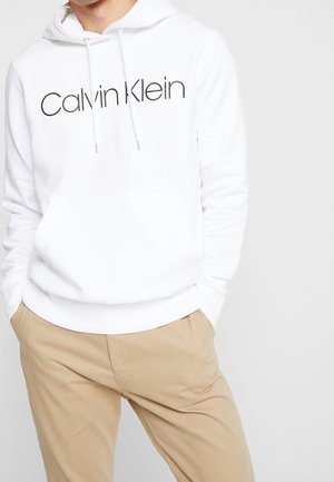 Felpa bianca con tasca a marsupio frontale, con il logo "Calvin Klein" nero. Un paio di pantaloni beige visibili in basso.