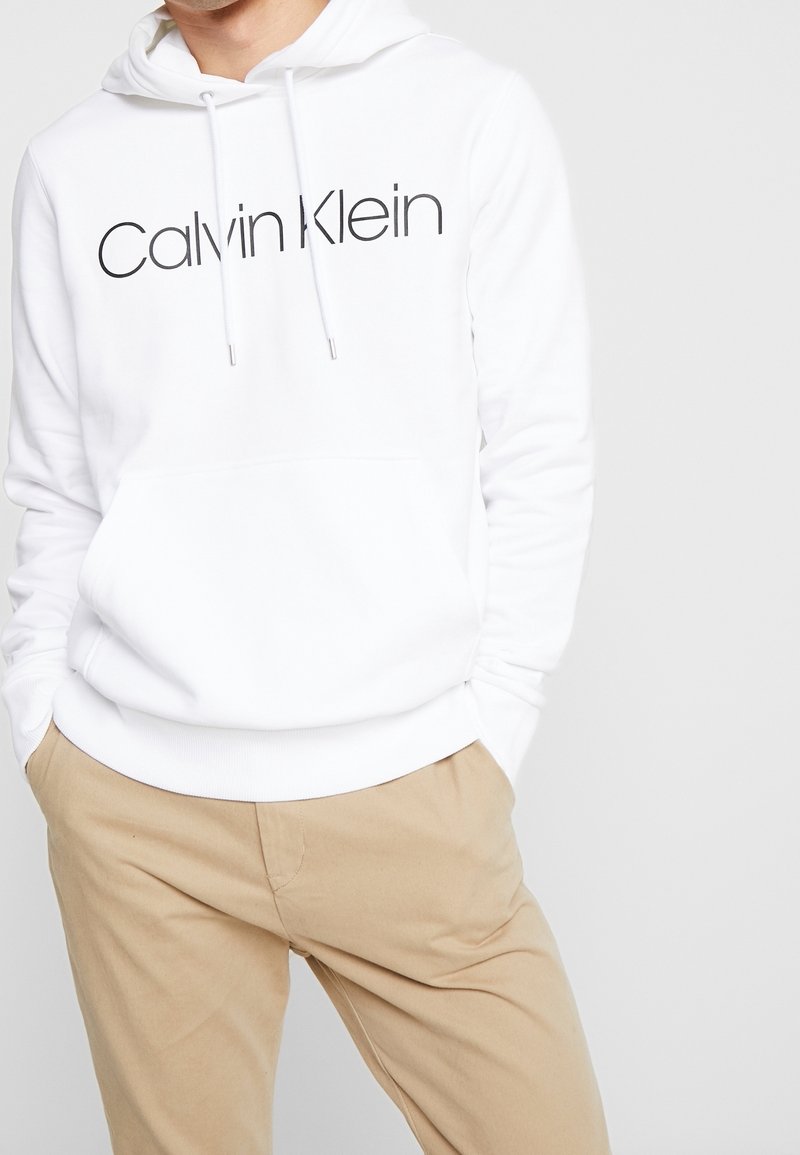 Sudadera blanca con un bolsillo canguro en la parte frontal, con un logo negro de "Calvin Klein". Pareja de pantalones beige visibles en la parte inferior.