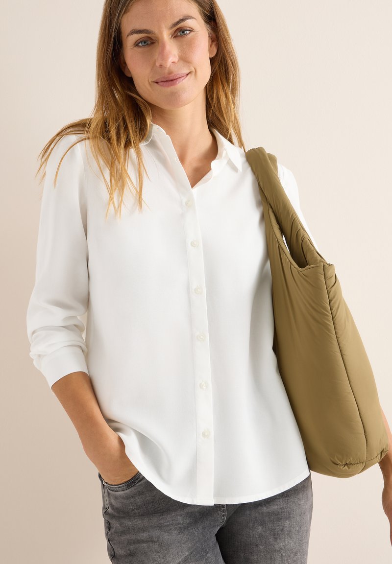 Chemise blanche à manches longues, texture lisse et ourlet arrondi, associée à un sac matelassé vert olive clair et un jean gris.