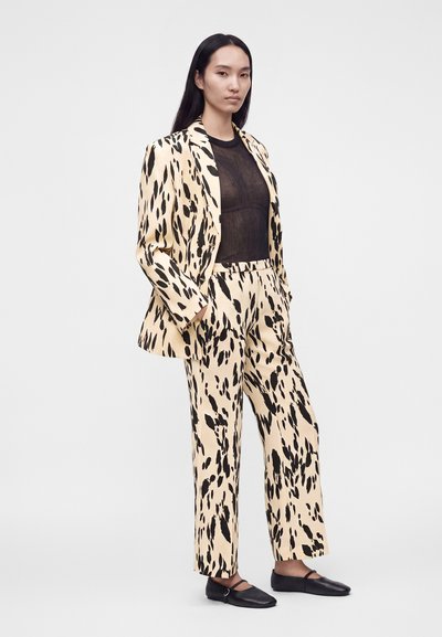 Femme aux longs cheveux foncés porte un costume beige à pois noirs avec pantalon assorti, un haut noir transparent et des chaussures plates noires, debout de profil.