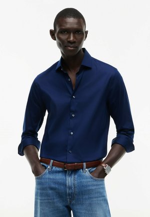 Uomo dalla pelle scura che indossa una camicia blu navy con bottoni, jeans azzurri, cintura marrone e orologio da polso, in piedi con le mani in tasca.