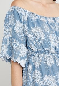 Sininen olkaimeton mekko, joka on valmistettu denimpainotteisesta kankaasta. Mekossa on valkoisia kukkakuosisia kohtia, röyhelöhihnat ja joustava vyötärö.