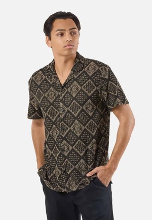 Jonge man met een zwart-beige kort overhemd met geometrische diamantpatronen, handen in de zakken, kijkt opzij.