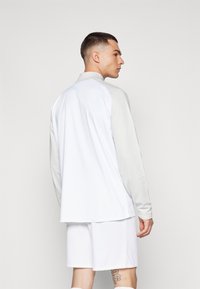 Outfit sportiv pentru bărbați, compus dintr-o bluză albă cu mânecă lungă și accente gri, alături de șorturi albe, realizate dintr-un material neted și ușor.