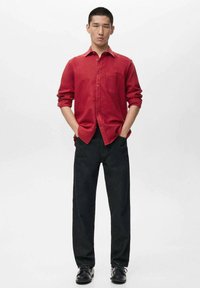 Rotes Hemd mit Knöpfen und langen Ärmeln, eine Brusttasche, kombiniert mit dunkler Jeanshose und schwarzen Schuhen, vor einem schlichten weißen Hintergrund.