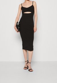 Černé šaty střední délky s přiléhavým střihem, s tenkými ramínky a výřezem vpředu. Modelka drží malou černou kabelku a má na sobě černé lodičky.