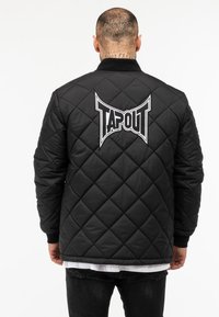 Čierna prešívaná bomber bunda s rebrovaným golierom, ktorá má na chrbte veľké biele logo "TAPOUT" a textúrované vypchávky.