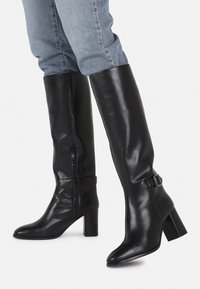 Bottes en cuir noir montant jusqu'au genou, avec une texture lisse, un bout pointu, un talon bloc, une fermeture éclair latérale et un détail de boucle à la cheville.