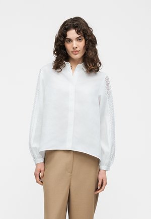 Kvinde iført en hvid bluse med blonder-detaljerede ærmer og beige bukser, stående mod en ensfarvet lys baggrund.