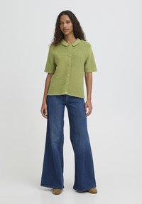 Chemise verte à manches courtes côtelées avec un col pointu et une fermeture boutonnée, associée à un jean large en denim bleu et des chaussures couleur tan.