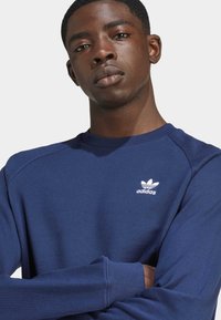 Navy zila džemperis ar apaļu kakla izgriezumu, ribotām piedurknēm un baltu Adidas logo uz krūtīm, izgatavots no mīksta kokvilnas materiāla.