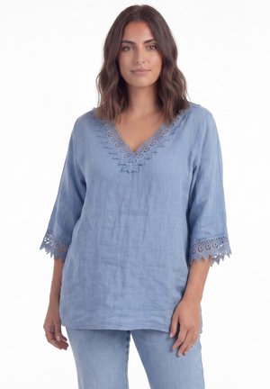 Femme aux cheveux bruns mi-longs portant un chemisier bleu clair avec bordure en dentelle au col en V et manches trois quarts, associé à un jean bleu clair.