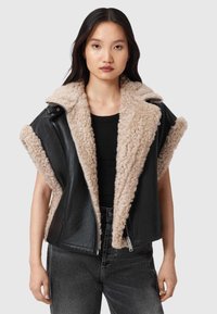 Juodos odos be rankovių striukė su smėlio spalvos sherpa apykakle ir pamušalu, turinti priekinį užtrauktuką ir tekstūruotas audinio detales.