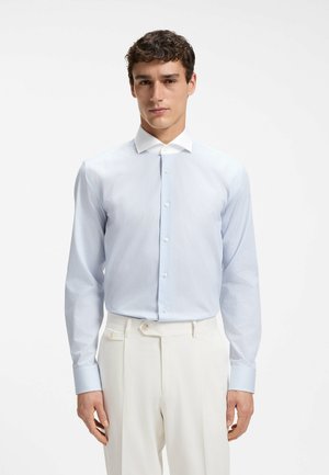 Man draagt lichtblauw gestreept overhemd met witte kraag en witte broek, staand tegen een effen achtergrond.