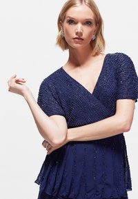 Top azul marino con cuentas, deescote en V y mangas cortas. Presenta un patrón de ondas texturizado y un corte holgado con dobladillo en capas.