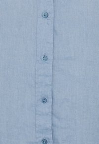 Ljusblå skjorta med knappar framtill, tillverkad av ett strukturerat tyg med sex mörkblå knappar. Inga synliga mönster eller detaljer.