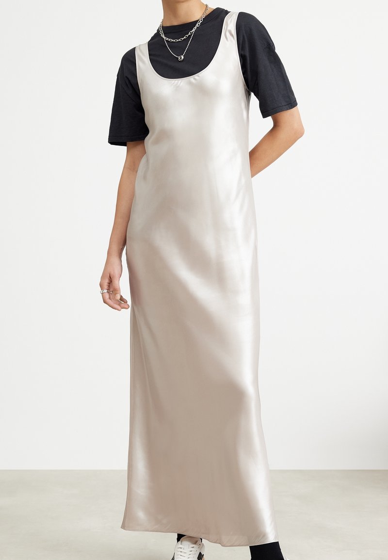 Personne portant une robe nuisette en satin argenté sans manches sur un t-shirt noir à manches courtes, accessoirisée de colliers et bagues argentés superposés.