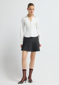 Jonge vrouw met slicked-back haar, gekleed in een witte strakke blouse met lange mouwen, een donkere geplooide rok, doorzichtige bordeauxrode sokken en metallic hakken.