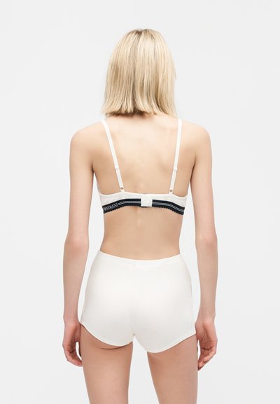 Lyshåret person med kort hår iført hvide shorts med høj talje og hvid bh med sort bånd, stående med ryggen mod en ensfarvet baggrund.