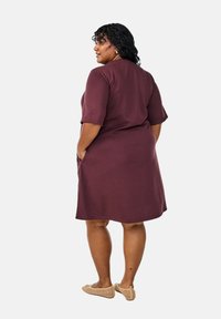 Maroon knee-length dress, short sleeves, relaxed fit, featuring side pockets. Smooth texture, plain design, paired with beige slip-on shoes.