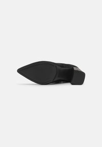Chaussure en suède noir avec un bout pointu, présentant un talon arrière texturé et une semelle plate en noir texturé. Lignes épurées et design minimaliste.