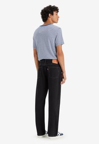 Młody mężczyzna w czarnych dżinsach, niebiesko-białej w pasy koszulce i niebieskich trampkach, stoi i patrzy w bok na białym tle.