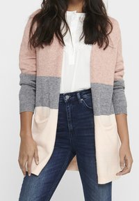 Žena nosící barevně rozdělený kardigan s růžovými, šedými a krémovými částmi přes bílé blouse a tmavě modré džíny.