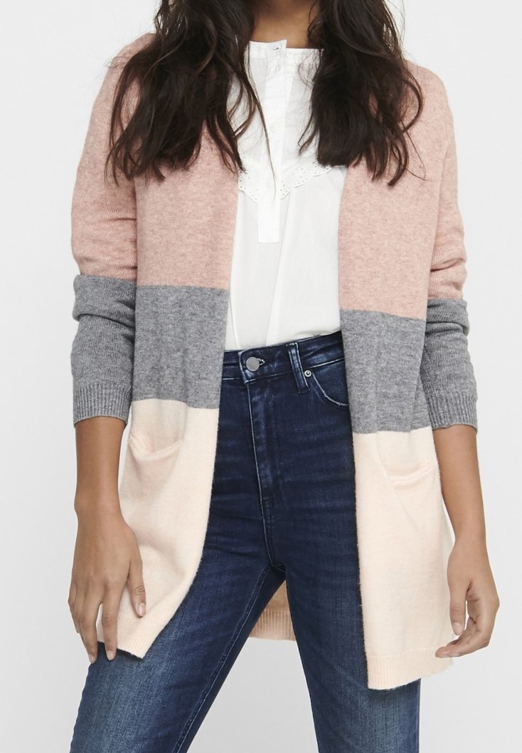 Mujer vistiendo un cárdigan de bloques de color con secciones en rosa, gris y crema sobre una blusa blanca y jeans azul oscuro.