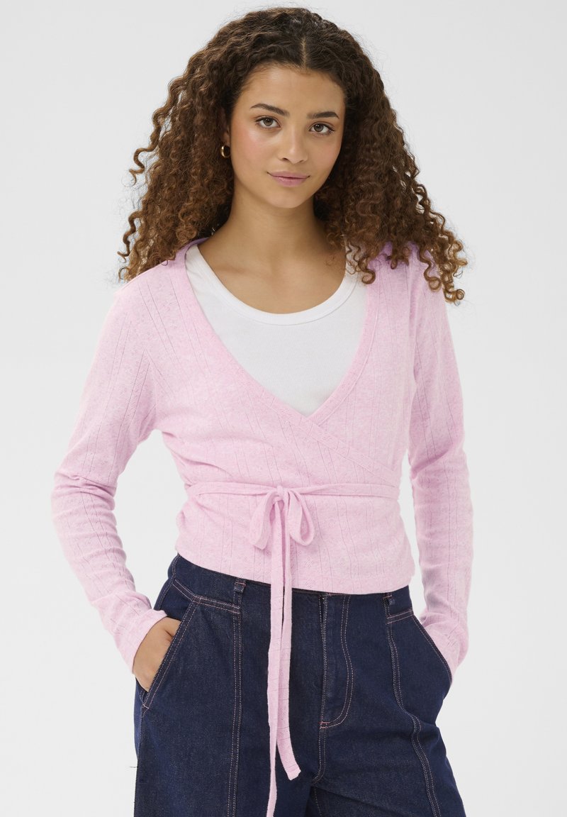 Fiatal nő göndör hajjal, világos rózsaszín, átvetős pulóvert visel fehér felső fölött, valamint sötétkék, magas derekú nadrágot, kezét a zsebében tartva.