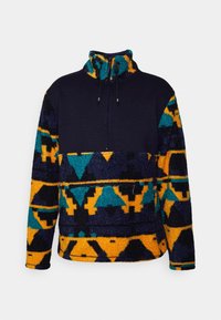Pull en polaire avec un haut bleu marine, motifs géométriques colorés en turquoise et orange sur la partie inférieure et les manches, équipé d'un col montant et d'une fermeture éclair.