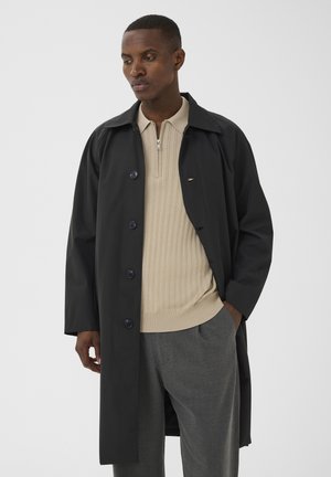 Homme portant un manteau noir boutonné par-dessus un pull beige à fermeture éclair et un pantalon gris, debout avec une main dans la poche, regardant vers le bas.