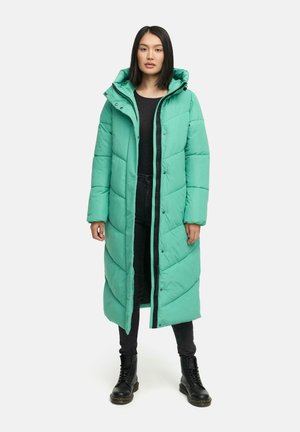 Doudoune turquoise avec capuche, présentant un design longline, une texture matelassée et une fermeture éclair à l'avant. Portée par-dessus un haut et un pantalon noirs.