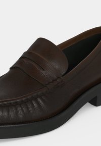 Mocassin penny en cuir marron foncé avec bout arrondi, bande décorative, coutures détaillées et semelle noire avec petit talon.