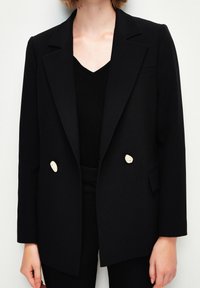 Černý propracovaný blazer s klopovým límcem, dvěma béžovými knoflíky, předními kapsami a strukturovaným střihem. Nosený přes černý top.