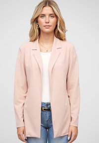 Lyserød blazer med lange ærmer, blød tekstur, åben front og enkle revers, båret over en hvid top og blå jeans.