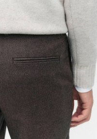 Brune teksturerede bukser med en smal baglomme. Stoffet har et subtilt diagonalt mønster, og en lysegrå sweater er delvist synlig.