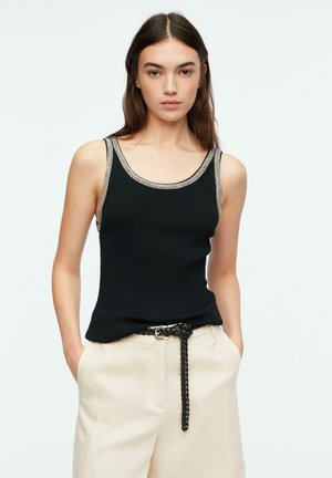 Giovane donna con lunghi capelli castani che indossa una canottiera nera con bordi beige, pantaloni beige e cintura nera intrecciata, mani in tasca.