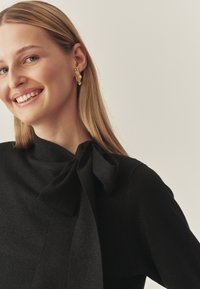 Femeie cu păr blond drept, purtând cercei mari din aur și un top negru cu un fundiță lângă gât, zâmbind ușor către stânga.