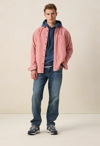 Roze corduroy shirt over een marineblauwe hoodie, gecombineerd met blauwe jeans en grijze sneakers. Het shirt heeft knopen en een borstzak.