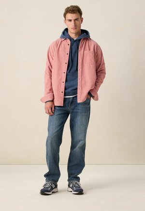 Chemise en velours côtelé rose sur un sweat à capuche navy, associée à un jean bleu et des baskets grises. La chemise possède des boutons et une poche poitrine.