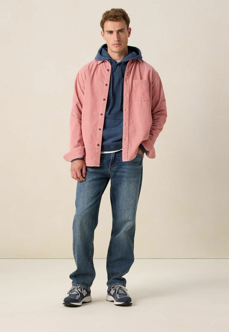 Roze corduroy shirt over een marineblauwe hoodie, gecombineerd met blauwe jeans en grijze sneakers. Het shirt heeft knopen en een borstzak.