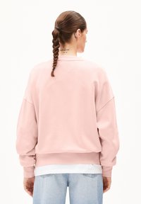 Ljusrosa sweatshirt med lös passform, sänkta axlar och långa ärmar. Buren över en vit lager topp med ribbad nederkant.