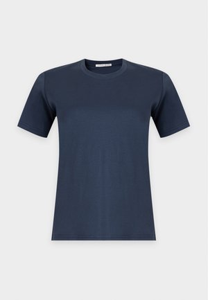 Kobieta o krótkich ciemnych włosach, ubrana w ciemnoniebieski t-shirt, szerokie fioletowe spodnie i czarne buty na spiczastym nosku, stojąca na jednolitym jasnym tle.