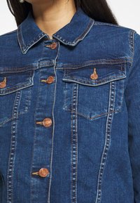 Kobieta w niebieskiej dżinsowej kurtce z miedzianymi guzikami i dwiema kieszeniami na piersi z zapięciem na guziki, widoczny kołnierz, długie ciemne włosy opadające na plecy.