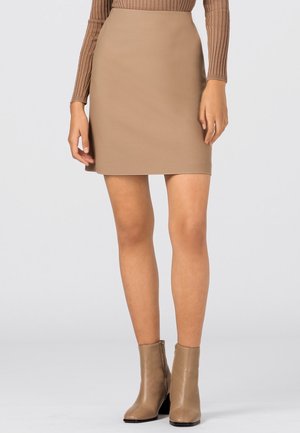 Kobieta w beżowej mini spódniczce, pasujących botkach za kostkę oraz prążkowanym brązowym topie z długim rękawem, stojąca na tle jednolitego, jasnego tła.