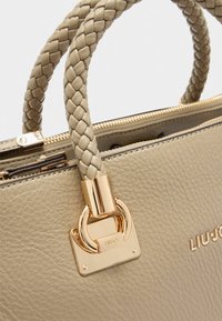 Sac en cuir beige avec des poignées tressées, des accents en métal doré et une fermeture éclair. Il comporte une plaque de logo et une surface texturée.