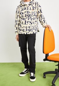 Beiger Kapuzenpullover mit dunkelblauen Textmustern, Reißverschlusstasche, schwarze Cargohose und schwarze Turnschuhe mit weißen Akzenten. Orangefarbener Stuhl in der Nähe.