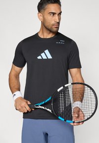 Melna sporta t-krekls ar gaiši zilu Adidas logotipu un tekstu. Modelis tur tenisa raketi ar melnu rokturi un zilām akcentiem.