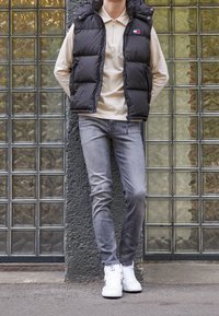Schwarze Steppweste über einem cremefarbenen Langarm-Polo, graue Skinny Jeans und weiße High-Top-Sneaker, stehend vor einer Glasbausteinwand.