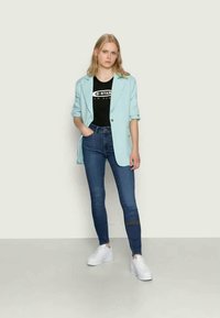 Jovem mulher a usar um blazer azul claro, t-shirt gráfica preta, jeans skinny azuis e ténis brancos de salto, de pé com uma mão no bolso.